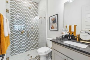 Modern bathroom in Painted Tree McKinney with a glass-enclosed chevron-tiled shower, white toilet, black-countertop vanity, and gold and orange accents—perfect for those seeking stylish homes for sale in McKinney, Texas.