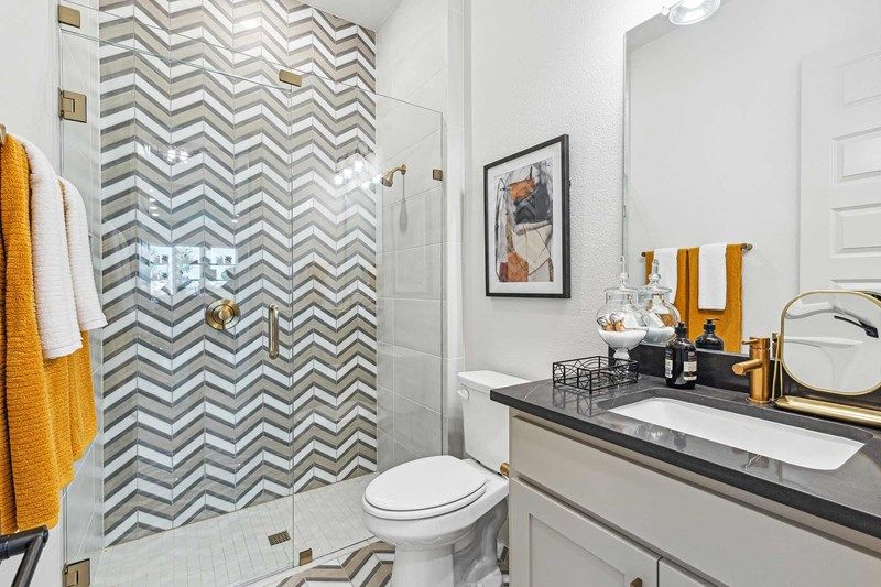 Modern bathroom in Painted Tree McKinney with a glass-enclosed chevron-tiled shower, white toilet, black-countertop vanity, and gold and orange accents—perfect for those seeking stylish homes for sale in McKinney, Texas.