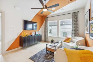 Modern living room with geometric orange accent wall, wall-mounted TV, black cabinet, armchair, patterned rug, and large windows—discover similar stylish spaces in Painted Tree McKinney while exploring homes for sale in McKinney, Texas.