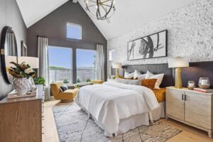 Contemporary bedroom with vaulted ceiling, large windows, white bedding, and modern artwork—an inviting retreat found in many Painted Tree homes for sale. A mustard-yellow accent chair by the window adds a pop of color to this stylish space.