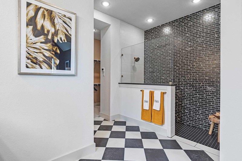 Modern bathroom with black-and-white checkered floor, black tiled shower, glass partition, two yellow towels, and a framed abstract artwork on a white wall—style that echoes the chic interiors found in Painted Tree McKinney homes for sale.