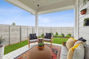 Covered patio with modern outdoor furniture, a round wicker table, decorative rug, and potted plants overlooks a fenced grassy yard under a blue sky—perfect for enjoying the Painted Tree McKinney lifestyle.