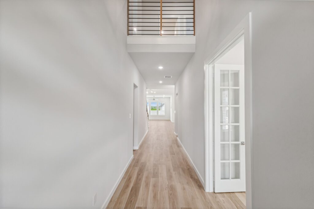 A bright hallway with light wood flooring, white walls, and a loft railing above leads to a windowed living area—discover elegant details like these in Painted Tree McKinney homes for sale in McKinney, Texas.