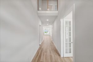 A bright hallway with light wood flooring, white walls, and a loft railing above leads to a windowed living area—discover elegant details like these in Painted Tree McKinney homes for sale in McKinney, Texas.