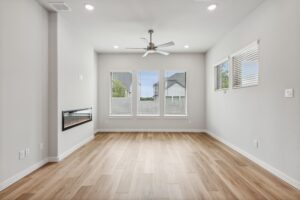A bright, empty living room in Painted Tree McKinney features light wood floors, white walls, large windows, a ceiling fan, and a modern wall-mounted fireplace—perfect for those seeking Painted Tree homes for sale.