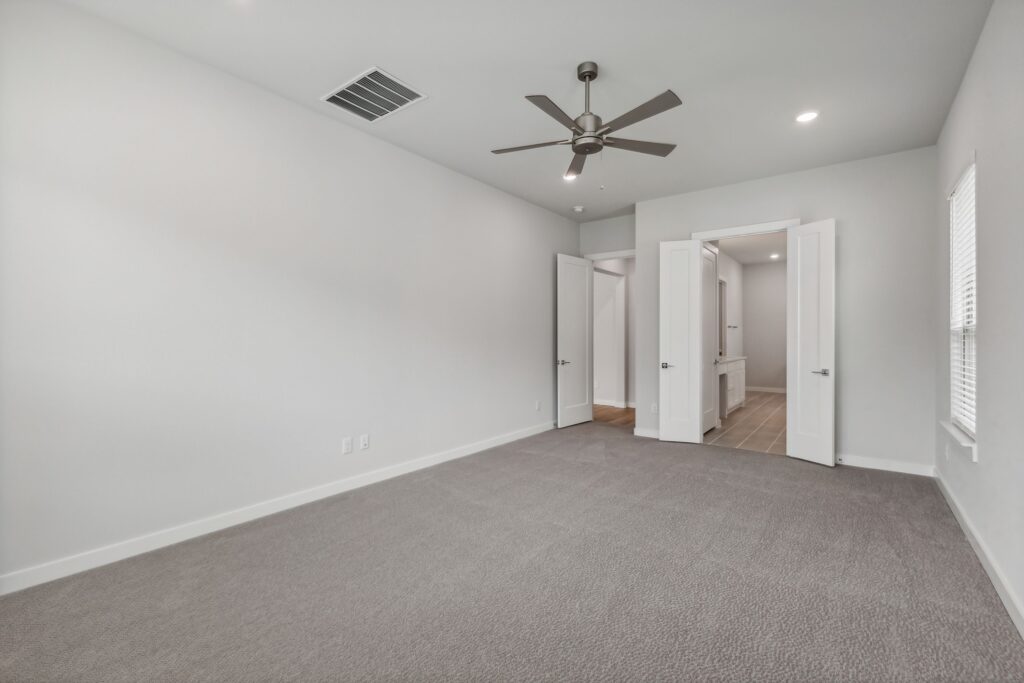 Empty, carpeted room with a ceiling fan, white walls, and two open doors leading to a hallway and bathroom. Natural light enters through a window on the right—one of many inviting spaces in homes for sale in McKinney, Texas.