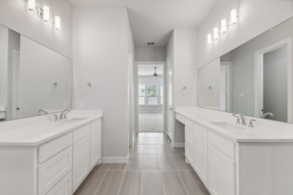 Modern bathroom with dual white vanities, large mirrors, and wall-mounted lights; an open doorway leads to a bright bedroom with a ceiling fan in one of the stunning homes for sale in McKinney, Texas.