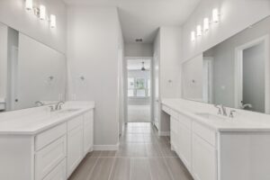 Modern bathroom with dual white vanities, large mirrors, and wall-mounted lights; an open doorway leads to a bright bedroom with a ceiling fan in one of the stunning homes for sale in McKinney, Texas.