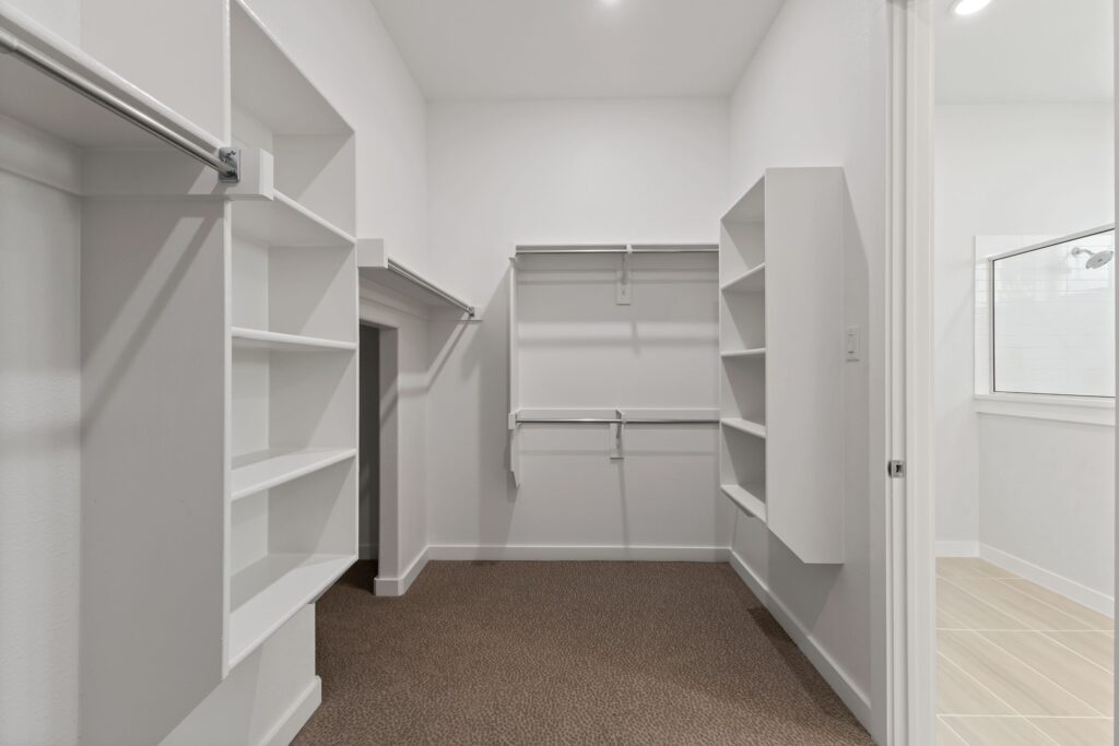 Empty walk-in closet with white shelves, hanging rods, and carpeted floor; adjacent bathroom with tile flooring visible through open door in one of the beautiful Painted Tree homes for sale in McKinney, Texas.