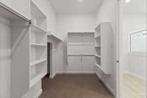 Empty walk-in closet with white shelves, hanging rods, and carpeted floor; adjacent bathroom with tile flooring visible through open door in one of the beautiful Painted Tree homes for sale in McKinney, Texas.