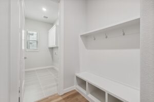 A white mudroom with built-in bench, cubbies, and wall hooks leads to a laundry area with tiled floor, window, and overhead cabinets—just one of the inviting spaces found in Painted Tree McKinney homes for sale.