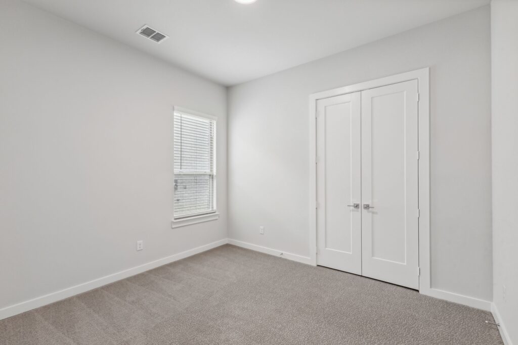 Bright and inviting, this empty room in Painted Tree TX features light gray carpet, white walls, a window with blinds, and double doors—an ideal canvas for your new beginning. Explore more Painted Tree homes for sale today!.