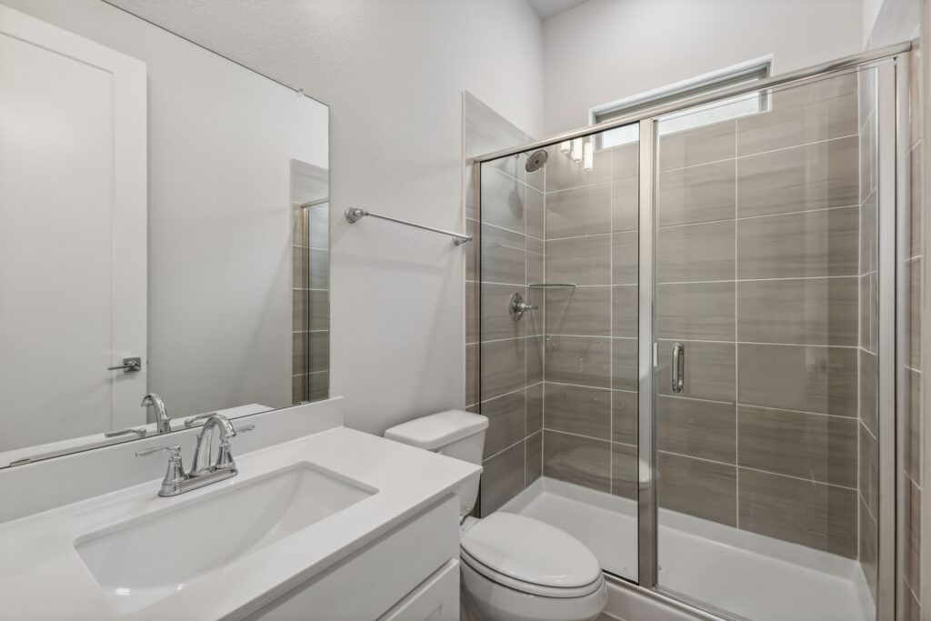 Modern bathroom in a Painted Tree McKinney home features a glass-enclosed shower, gray tile walls, white vanity with sink, large wall mirror, and a sleek white toilet—perfect for those seeking homes for sale in McKinney Texas.