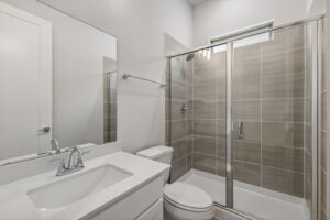 Modern bathroom in a Painted Tree McKinney home features a glass-enclosed shower, gray tile walls, white vanity with sink, large wall mirror, and a sleek white toilet—perfect for those seeking homes for sale in McKinney Texas.