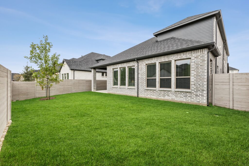 A backyard with a well-maintained green lawn, young tree, wooden fence, and a brick house with multiple windows under a partly cloudy sky—perfect for those seeking homes for sale in McKinney, Texas.