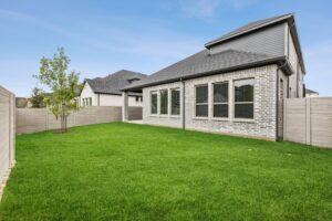 A backyard with a well-maintained green lawn, young tree, wooden fence, and a brick house with multiple windows under a partly cloudy sky—perfect for those seeking homes for sale in McKinney, Texas.