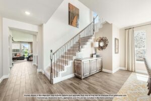 A bright entryway features a staircase with metal railings, a wooden sideboard, and a round mirror; adjacent rooms are visible with large windows and light wood flooring—perfect for those seeking homes for sale in McKinney, Texas.