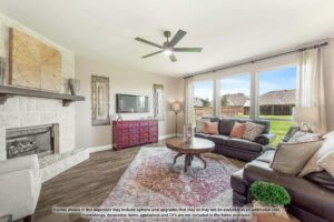 Bright living room with a stone fireplace, wall-mounted TV, patterned rug, and brown sofas. Large windows overlook a fenced backyard in Painted Tree McKinney. Text disclaimer appears at the bottom of the image.