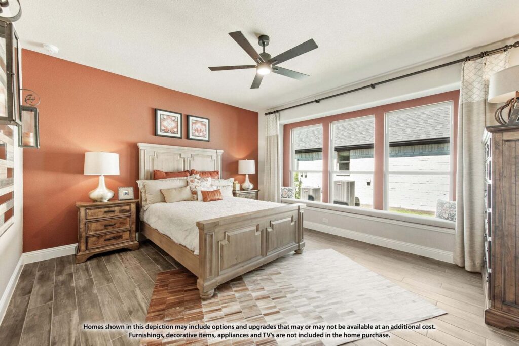 A bedroom in Painted Tree TX features a wooden bed, nightstands, lamps, wall art, a ceiling fan, large windows with curtains, and wood-look tile flooring—perfect for those seeking homes for sale in McKinney Texas.