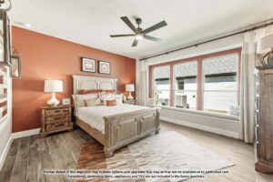 A bedroom in Painted Tree TX features a wooden bed, nightstands, lamps, wall art, a ceiling fan, large windows with curtains, and wood-look tile flooring—perfect for those seeking homes for sale in McKinney Texas.
