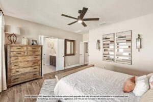 Bedroom with a wooden dresser, bed with white bedding, ceiling fan, wall art, and an open door leading to a bathroom. Natural light enters from the left—ideal for those seeking Painted Tree McKinney homes for sale.
