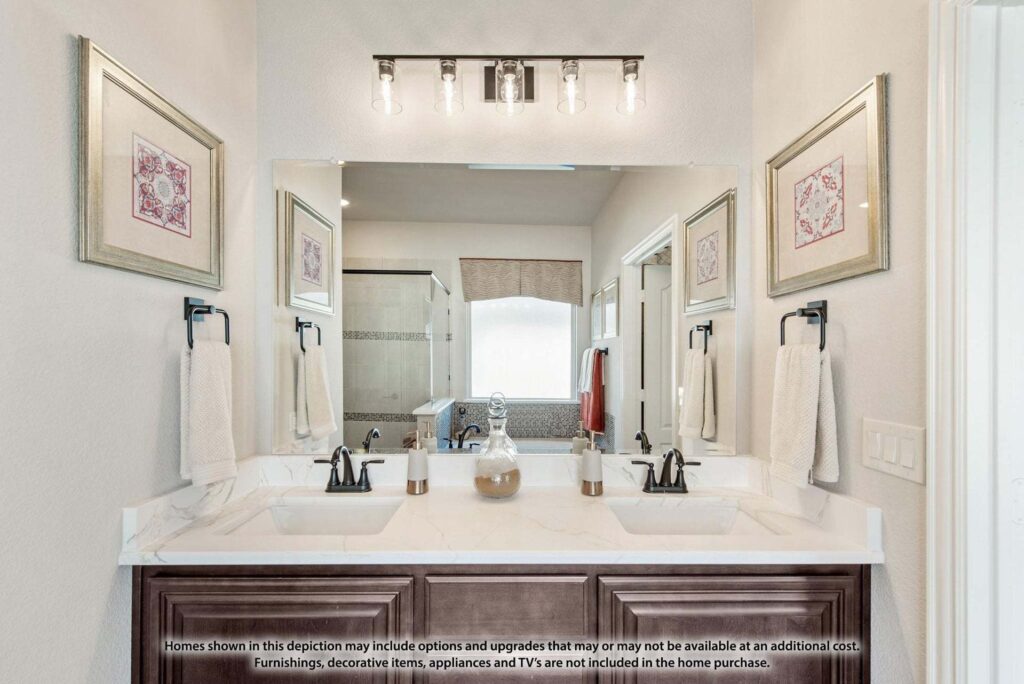 Modern bathroom with double sink vanity, wall-mounted mirrors, light fixture, towel racks, and glass-enclosed shower in the background. Discover elegant finishes like this in Painted Tree McKinney homes for sale. Text disclaimer visible on the sink counter.