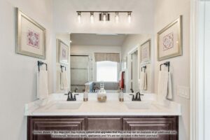 Modern bathroom with double sink vanity, wall-mounted mirrors, light fixture, towel racks, and glass-enclosed shower in the background. Discover elegant finishes like this in Painted Tree McKinney homes for sale. Text disclaimer visible on the sink counter.