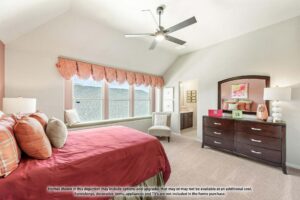 A bedroom with a red bedspread, pillows, dresser with mirror, bedside lamps, and large windows with valances—just one inviting space you’ll find in Painted Tree McKinney homes for sale. A doorway leads to an adjoining bathroom.