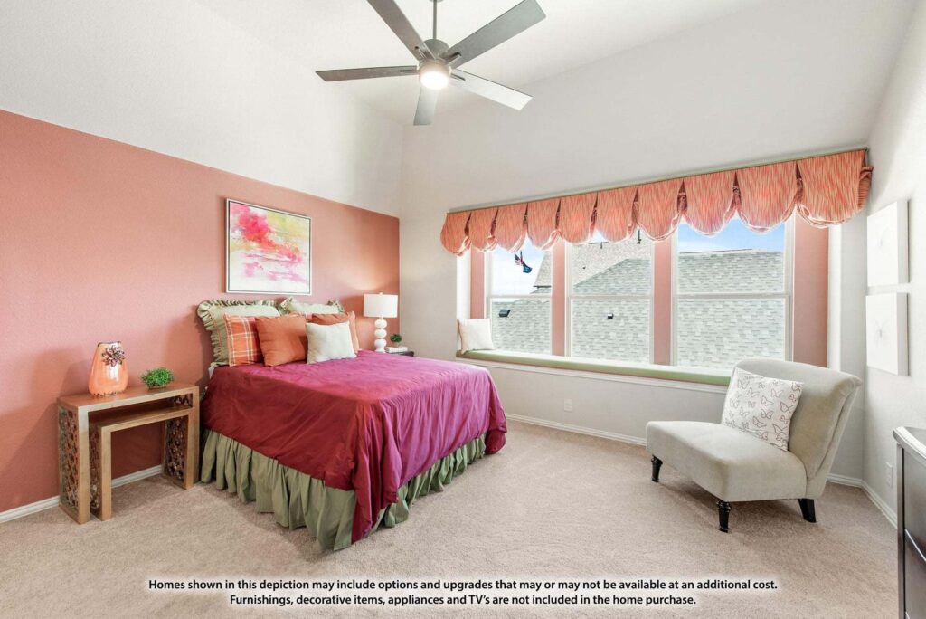 Bedroom in Painted Tree TX features a red accent wall, bed with red and green bedding, bedside table, upholstered chair, large windows with valance, and ceiling fan—one of the stylish options among homes for sale in McKinney Texas.