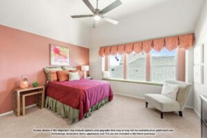 Bedroom in Painted Tree TX features a red accent wall, bed with red and green bedding, bedside table, upholstered chair, large windows with valance, and ceiling fan—one of the stylish options among homes for sale in McKinney Texas.
