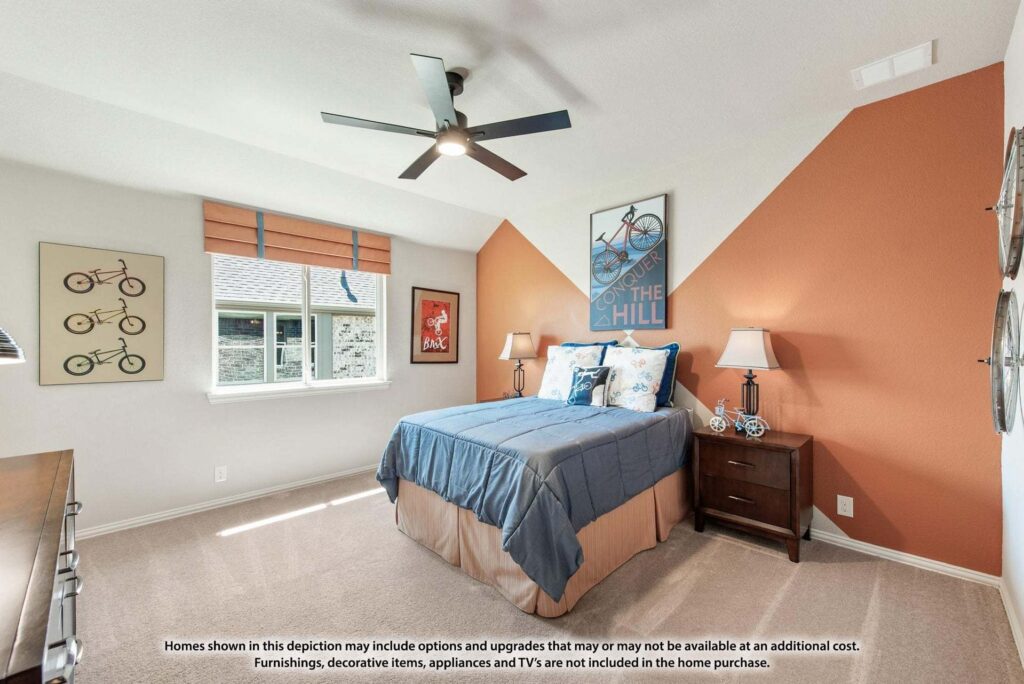 A neatly arranged bedroom in Painted Tree TX features a blue bed, orange accent wall, ceiling fan, nightstand, and framed wall art. A large window fills the space with natural light—perfect for those seeking homes for sale in McKinney Texas.