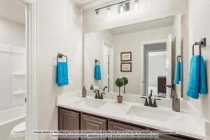 Modern bathroom with double sinks, marble countertop, large mirror, blue hand towels, and a glimpse of a shower-tub combo—one of the highlights found in many Painted Tree homes for sale in McKinney, Texas.