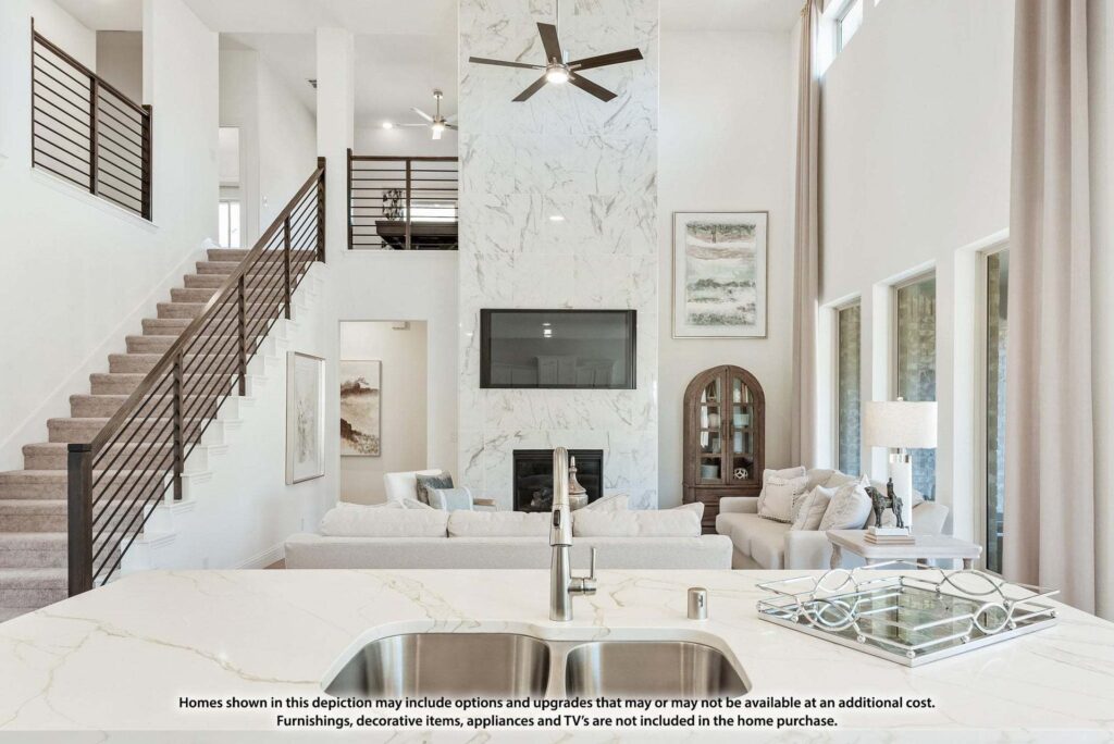Modern living room with high ceilings, neutral decor, fireplace with TV above, stairway, and large windows—all viewed from a kitchen island with a sink—offers stunning style in sought-after Painted Tree TX homes for sale.