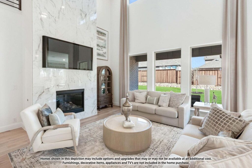 Modern living room with tall ceilings, marble fireplace, mounted TV, neutral-toned sofas, large windows, and a round coffee table—discover stylish comfort in Painted Tree TX; notice about furnishings at the bottom.