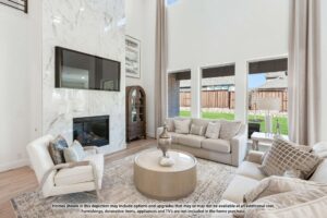 Modern living room with tall ceilings, marble fireplace, mounted TV, neutral-toned sofas, large windows, and a round coffee table—discover stylish comfort in Painted Tree TX; notice about furnishings at the bottom.