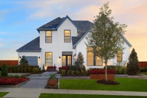 Modern two-story house with white exterior, large windows, landscaped yard, and driveway under a partly cloudy sky at sunset in Painted Tree McKinney—explore beautiful Painted Tree homes for sale in TX.