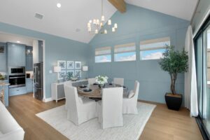 A modern dining room in Painted Tree TX features light blue walls, a round table set for four, white slipcovered chairs, a chandelier, large windows, and a potted plant in the corner. Discover homes for sale in McKinney Texas with stunning interiors like this.