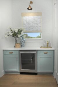 A small home bar with light blue cabinets, a mini fridge, glass decanters on a tray, decorative vases, and white flowers—perfect for entertaining in one of the beautiful Painted Tree homes for sale. A window with a woven shade sits above the counter.