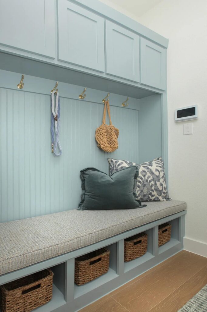 A built-in light blue mudroom bench with cushion, throw pillows, wall hooks holding a bag and leash, and four wicker baskets in cubbies below—perfect for homes for sale in McKinney Texas like those in Painted Tree McKinney.