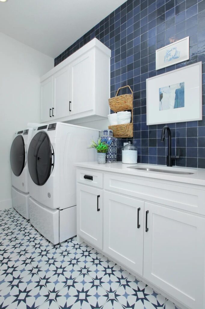 Modern laundry room in Painted Tree McKinney with white cabinets, blue patterned tile floor, stacked washer and dryer, countertop, sink, and stylish baskets and artwork—perfect for those seeking homes for sale in McKinney, Texas.