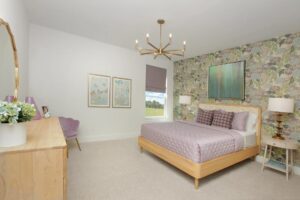 Modern bedroom with a light wood bed, lavender bedding, floral wallpaper accent wall, chandelier, dresser, and framed art. A window reveals greenery—capturing the serene style found in Painted Tree McKinney homes for sale.
