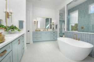 Modern bathroom with light blue cabinets, a freestanding white bathtub, gold fixtures, dual sinks, and blue tiled shower—an inviting space often found in painted tree homes for sale or sought-after homes for sale in McKinney Texas.