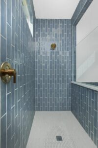 A stylish shower features vertical blue rectangular tiles, a brass showerhead and handle, a small window, and a white tiled floor—an inviting detail in many Painted Tree McKinney homes for sale.