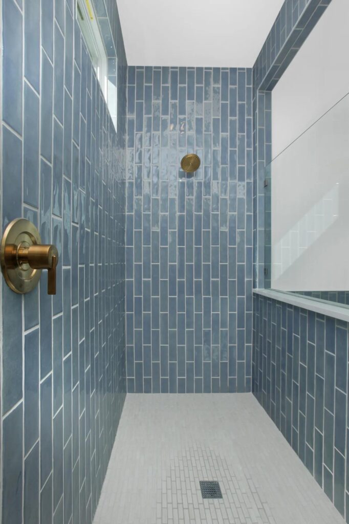 A stylish shower features vertical blue rectangular tiles, a brass showerhead and handle, a small window, and a white tiled floor—an inviting detail in many Painted Tree McKinney homes for sale.