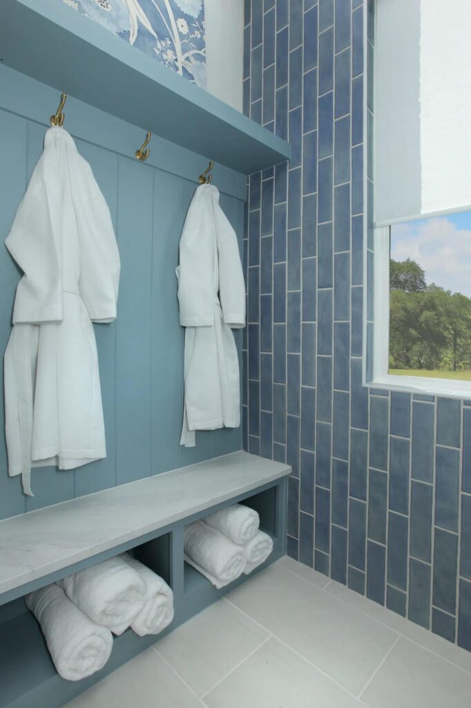 Two white bathrobes hang on hooks above a bench with shelves holding rolled white towels; blue tiles and a large window add style—perfect inspiration for homes for sale in McKinney, Texas.