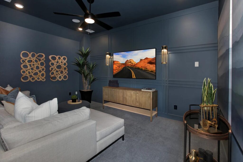 Modern home theater room with dark blue walls, gray sectional sofa, wall-mounted TV, wooden console, plants, and decorative wall art—perfect for relaxing in one of the stunning homes for sale in McKinney, Texas.