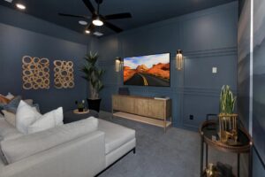 Modern home theater room with dark blue walls, gray sectional sofa, wall-mounted TV, wooden console, plants, and decorative wall art—perfect for relaxing in one of the stunning homes for sale in McKinney, Texas.