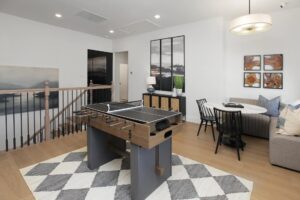 A game room in Painted Tree TX featuring a table tennis setup atop a foosball table, a round dining table with chairs, modern wall art, and a wooden-railed staircase—perfect for those seeking homes for sale in McKinney Texas.