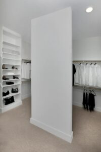 A walk-in closet with shelves of shoes and folded clothes on the left, and hanging black and white clothing on racks along the walls. A large white partition stands in the center—luxury you’ll find in many painted tree homes for sale.