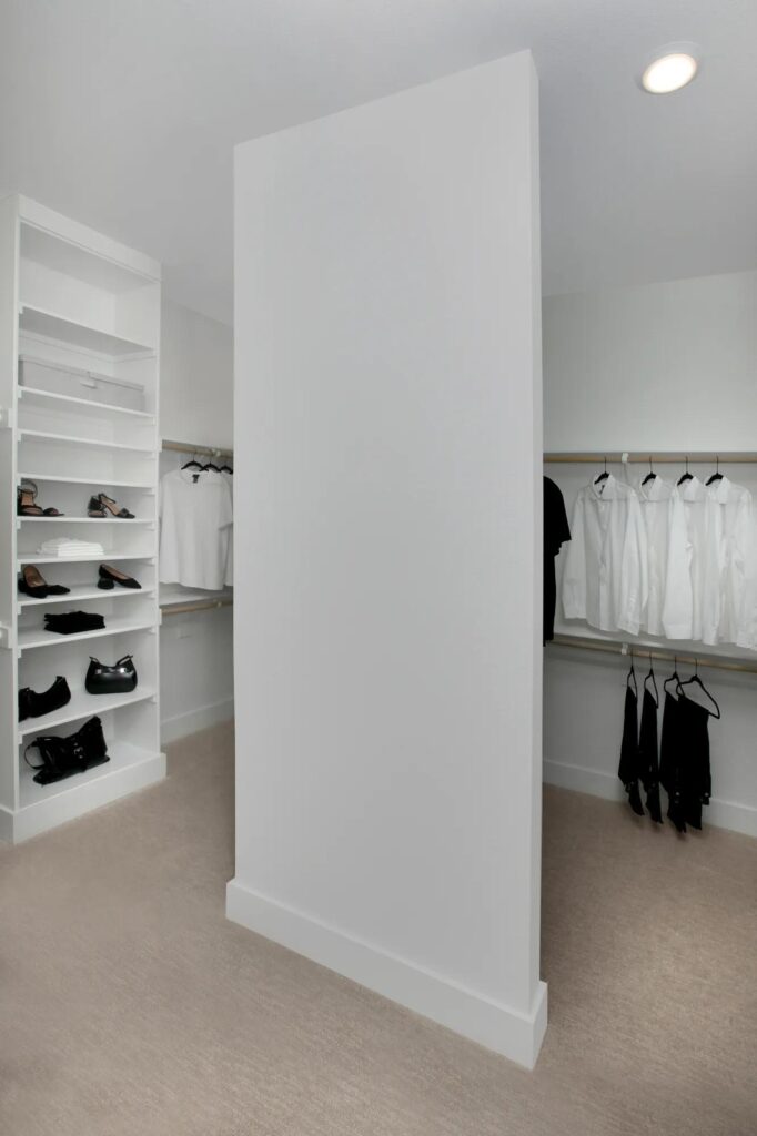 A walk-in closet with shelves of shoes and folded clothes on the left, and hanging black and white clothing on racks along the walls. A large white partition stands in the center—luxury you’ll find in many painted tree homes for sale.